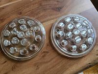 Top view of two circular clear glass flower frogs with 16 holes each arranged in concentric circles, placed on a wooden surface.