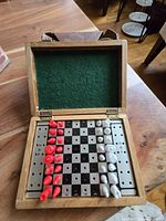 Wooden travel chess set opened to show chessboard and red and white pieces placed on it. Board has holes to secure pieces during travel, felt lining inside lid.