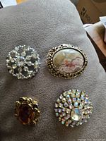 Top view of four vintage brooches on gray surface, showing details of rhinestones, cameo design, and gold-tone frames.
