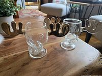Two clear glass egg nog glasses each with gold-tone moose antlers and skull shaped lower section, photographed on wooden table in living room setting.