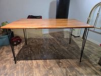 Photo showing full view of MCM style dining table with visible steel 3-pin legs and oak laminated tabletop against a wood floor background.