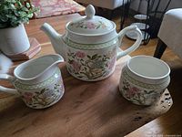 Photo showing teapot, creamer, and open sugar bowl with floral design on wooden table.