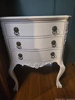 Front view of the white painted Gibbard nightstand with three drawers and brass handles, showing curved cabriole legs and ornate scrollwork on the apron.