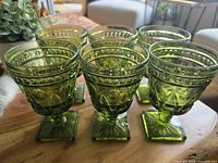 Front and side angle view showing six medium green goblets with faceted stems and square bases arranged on a wooden surface, highlighting the rich green color and pressed details.