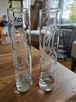 Front view of two cylindrical clear glass vases with etched indigenous-style frog and whale/bird designs standing side by side on a wooden table.