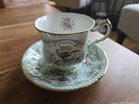 Tea cup and saucer set displayed on wooden table showing bird and floral pattern with gold trim and scalloped edges