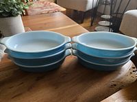 Six vintage light blue Pyrex oval dishes stacked in two piles of three on a wooden surface, showing their shape and size.