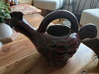 Side angle showing redware rooster teapot with loop handle over the top and integrated spout in beak area. Glazed brown finish with rustic folk-art style.