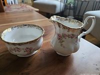 Front angle photo of vintage Royal Albert Dimity Rose small creamer and open sugar bowl on wooden table showing floral pattern and gold trim.