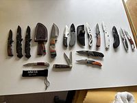 Overview of all 16 knives arranged on a white surface showing variety in blade length, handle design, and locking mechanism presence.