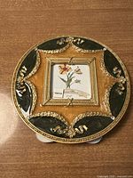 Front of the round pewter frame showing gold and green enamel pattern with floral and scroll motifs, holding a 2x2 inch photo insert of a flower illustration and labeled 'Pewter'.