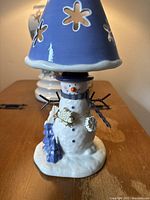 Snowman ceramic candle holder with blue top hat, scarf, and base. Snowflake-shaped cutouts on removable blue cone top. White and blue color scheme with detailed figurine elements including wreath and Christmas tree.