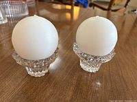 Two clear glass fluted base candle holders each holding a white globe candle, placed on a wooden surface.