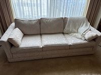 Front view of cream fabric sofa with three back cushions, floral pattern visible on fabric, and throw blanket on right arm.