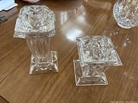 Two crystal candle holders of different heights with square bases and geometric cuts, placed on a wooden table. One holder shows visible chip on the top edges.