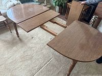 Round wooden dining table partly extended with leaf, showing turned legs and warm brown finish on carpet.