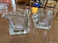 Pair of clear glass dishes with floral etching on wooden surface. Both have small handles and flower/starburst designs etched in glass.