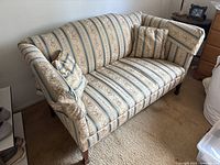 Front angle side view of the beige and blue striped floral fabric couch showing texture and wooden legs. Two matching striped throw pillows are placed on the couch.