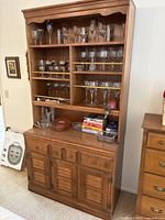 Front view of wooden cabinet displaying upper shelves with various glassware including wine glasses and goblets, books and other small items on counter space, and lower section with drawer and three louvered doors.