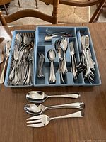 Set of stainless steel flatware including forks, spoons, knives organized in blue plastic storage tray and loose serving utensils on table.