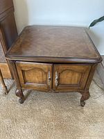 Full view of the wooden end table showing the double doors, decorative handles, and carved legs on carpeted floor.