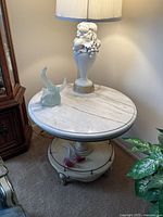 Full view of the vintage cream-colored marble top table with a decorative lamp and glass figurine on top, showcasing the round marble surface and brass accents.
