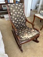 Front view of vintage wooden rocking chair upholstered in dark brown fabric with beige floral pattern. Visible damage worn on front seat area.
