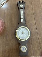 Full view of the wall-mounted weather instrument showing thermometer, barometer, and hygrometer on wooden backing.
