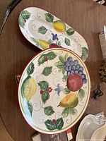 Two plates, one round and one oval, placed on a wooden table showing colorful painted fruit and leaf patterns with red border.