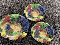 Three Fitz and Floyd porcelain plates featuring roosters and vegetables, with vibrant colors and sculptural detailing, shown on a dark carpet background.