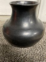 Front and side angle view showing the wide rounded body, narrow neck, and flared rim of the blackware pottery bowl placed on carpet.