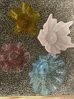 Four decorative glass bowls displayed on carpet: mustard yellow, soft pink, light blue, and deep rose colors with various ruffled and textured shapes.