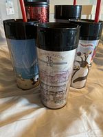 Five Starbucks travel tumblers with black lids and straws, displayed upright on a cloth surface. Designs include pandas, a 25th anniversary commemorative print, and scenic images.