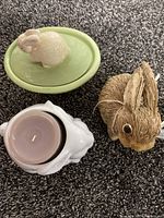Green oval ceramic rabbit container with lid, white ceramic rabbit candle holder with pink candle, brown straw rabbit figure