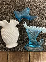 Top-down view of three glass items including a white hobnail milk glass vase with ruffled top, blue hobnail textured cornucopia vase with ruffled edge, and a blue glass bird figurine, all placed on a dark carpet.