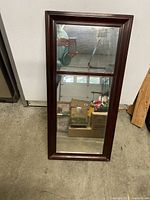 Front view of medium-sized rectangular mirror with dark wooden frame against a plain wall. Mirror is divided into an upper and lower section by wood trim. Reflects a person taking photo and storage items in background.