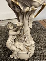 Side view showing the sculpted female figure climbing the vase with detailed floral designs and gilded accents on the off-white porcelain.