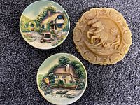 Two round ceramic decorative plates with English cottage scenes and one round wax carved plate depicting a mounted knight.