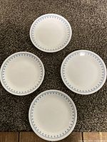 Four Corelle dinner plates arranged on a carpet, showing white base with blue floral/star pattern on rim