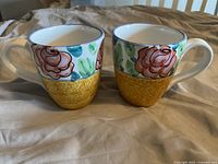 Two Starbucks ceramic mugs with floral and gold-speckled designs, showing rim chips, placed side by side on a fabric surface