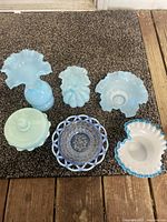 Six vintage Fenton glass pieces arranged on brown carpet, including vases and bowls in light blue and opalescent glass.