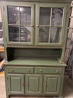 Full view of green wooden hutch showing upper glass-paneled display doors and lower drawer and cabinet storage.