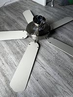 Ceiling fan shown on the floor with exposed wiring and five white blades attached to a brushed nickel motor housing.