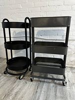 Photo showing two black metal salon trolleys side by side with a white brick background on wooden floor: the left has three round solid trays, the right has three rectangular mesh trays.
