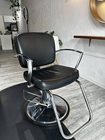 Front view of Pibbs Pisa salon styling chair showing black vinyl seat and backrest, chrome arms, hydraulic pump and round base on salon floor.