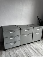 Photo showing three gray metal filing cabinets placed side by side with three drawers each, locks on one drawer per cabinet, all on caster wheels, standing on wood flooring against a white wall.