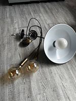 Three pendant lights laid out on a wood-look floor board; one with a large white metal shade and white bulb, and two with exposed large warm Edison bulbs attached to thin black and brass rods.
