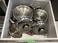 Top-down view showing various pieces of stainless steel cookware including frying pans, pots, and lids inside a storage drawer.