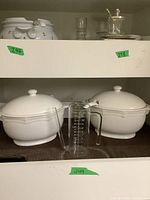 Three white ceramic soup bowls with domed lids, placed on a shelf. Two bowls are side by side with a glass measuring cup in front.