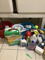 Photo showing two large buckets, boxes of gloves and dusters, several cleaning liquid bottles, sponges and mop heads grouped on the floor near a laundry setup.